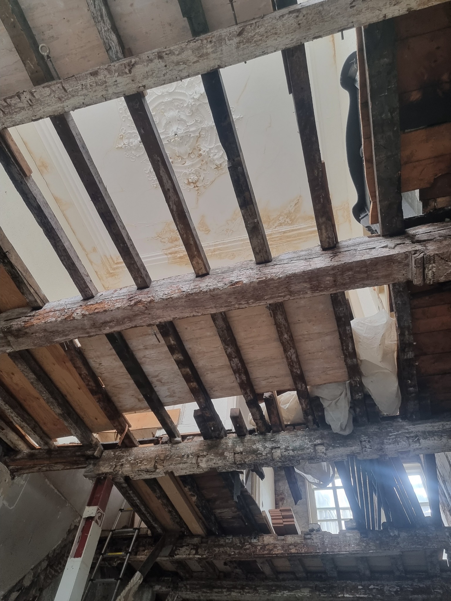 Demontaż stropu drewnianego z widocznymi belkami i fragmentami starego, zdobionego sufitu z plamami wilgoci, ukazujący etap prac przygotowawczych do remontu, z oknem w tle.