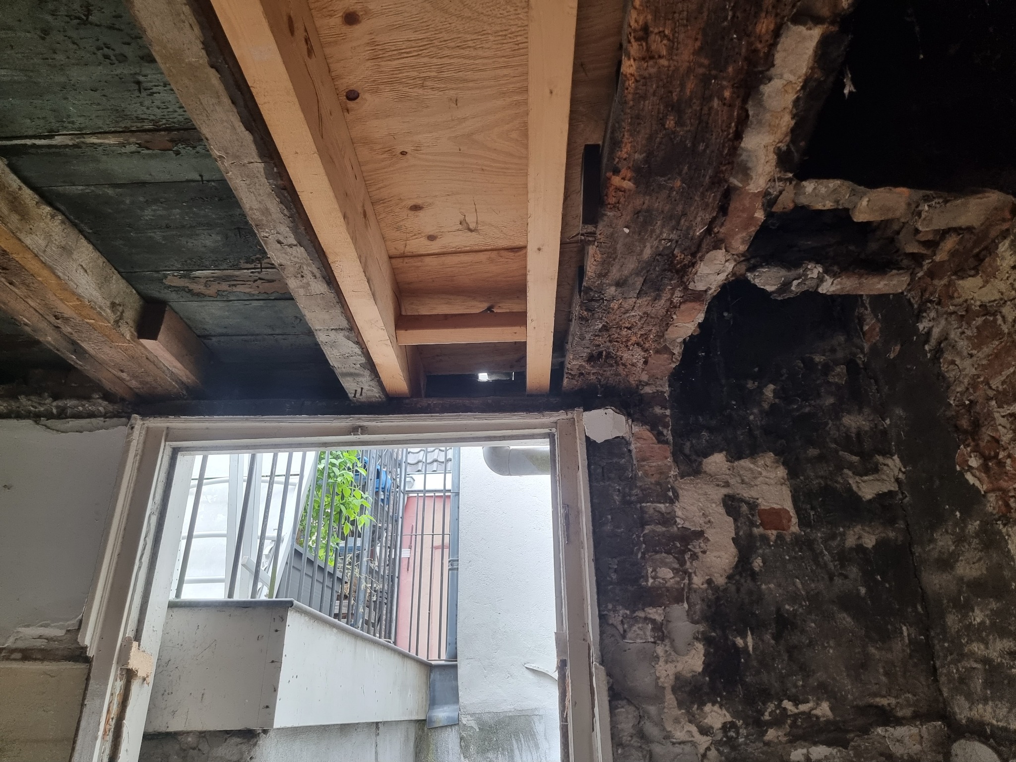 Fragment remontowanego pomieszczenia z odsłoniętym drewnianym stropem, fragmentem muru z cegły i widokiem na zewnątrz przez drzwi.