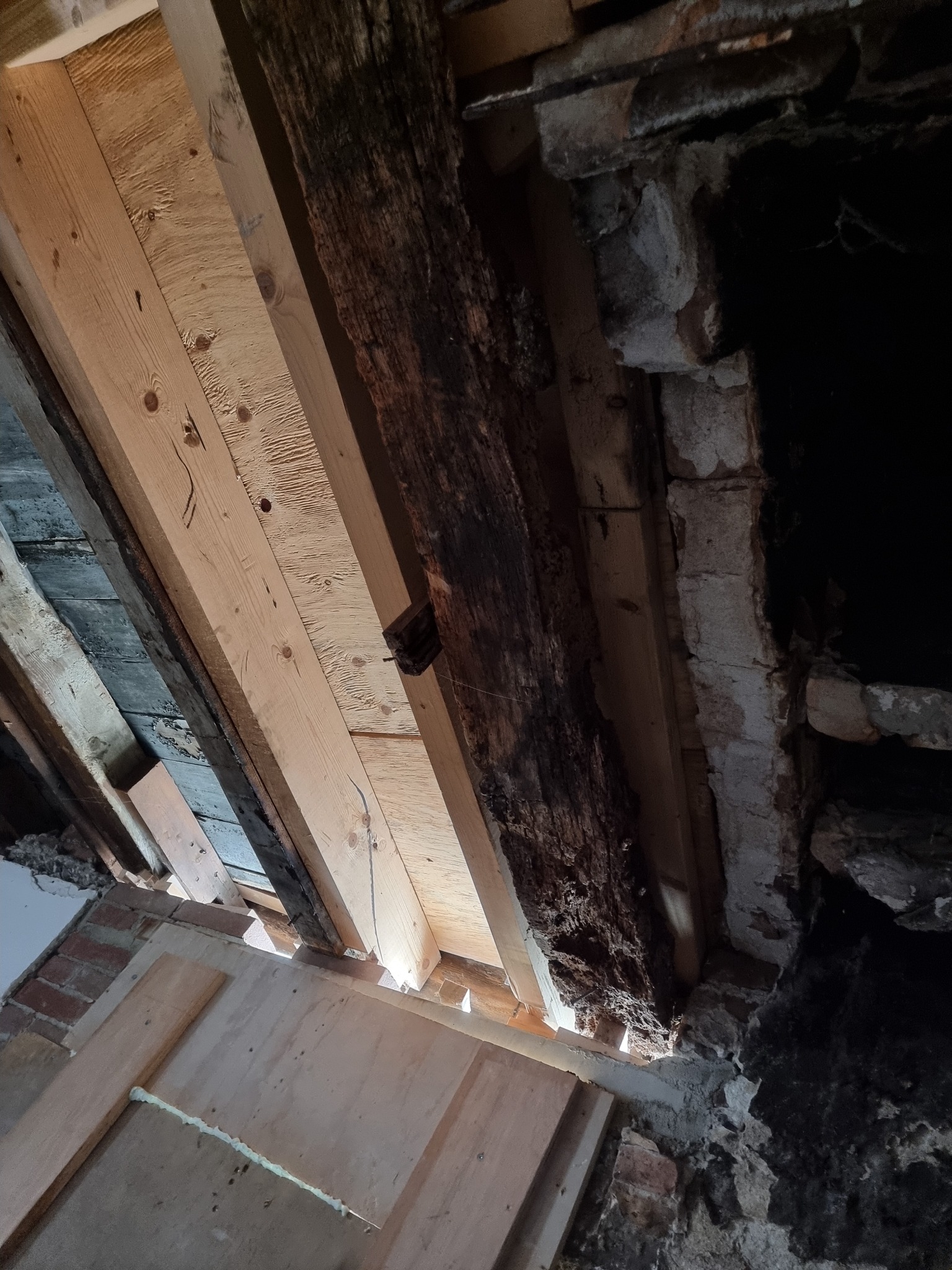Fragment konstrukcji stropu z drewnianymi belkami, nowymi i starymi, oraz fragment muru z cegły i kamienia w trakcie prac remontowych.