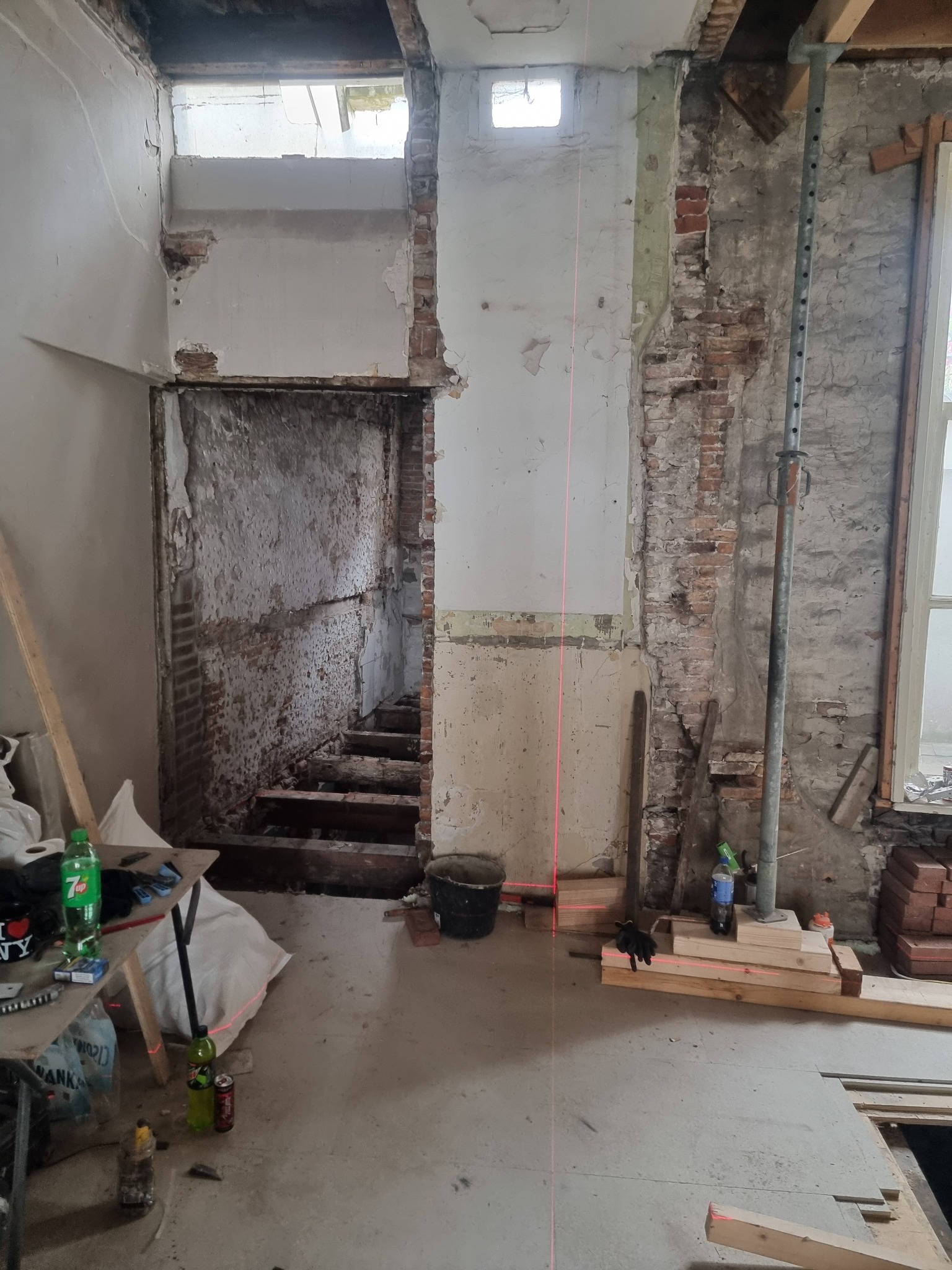Wnętrze w trakcie remontu: odsłonięte cegły, resztki tynku, drewniane schody w głębi, laser krzyżowy wyznaczający pion, podpory stropu i narzędzia budowlane.