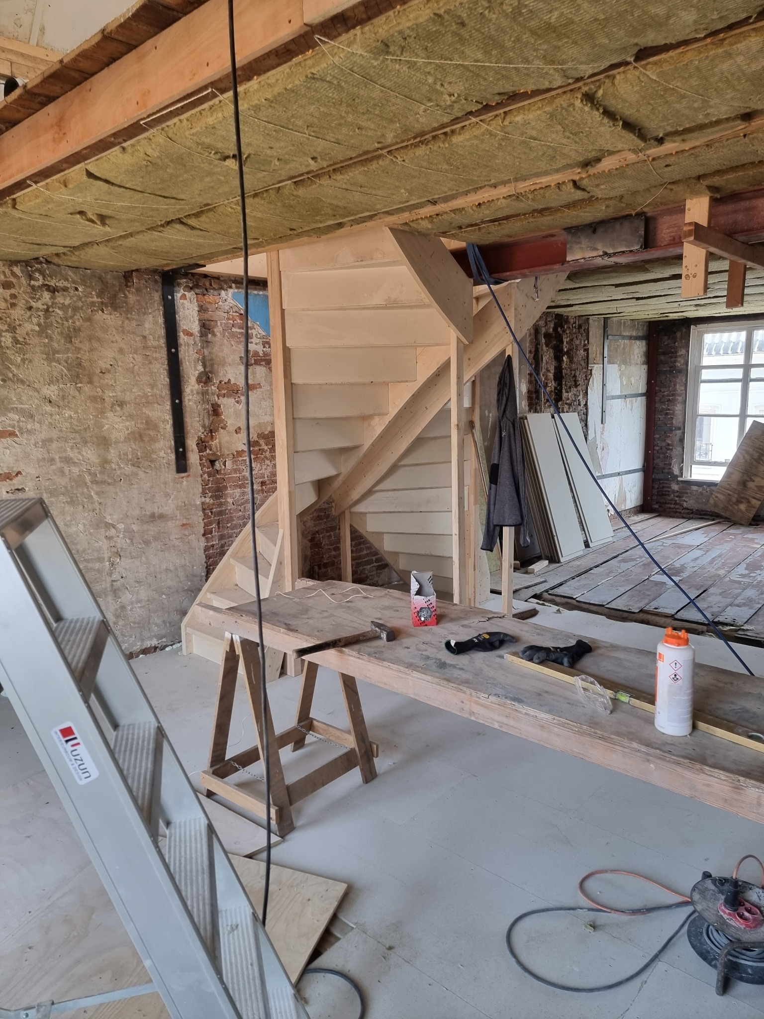 Wnętrze w trakcie renowacji z surową drewnianą konstrukcją schodów, widoczne ocieplenie sufitu wełną mineralną, fragment ściany z odsłoniętą czerwoną cegłą, drabina aluminiowa i stół roboczy...