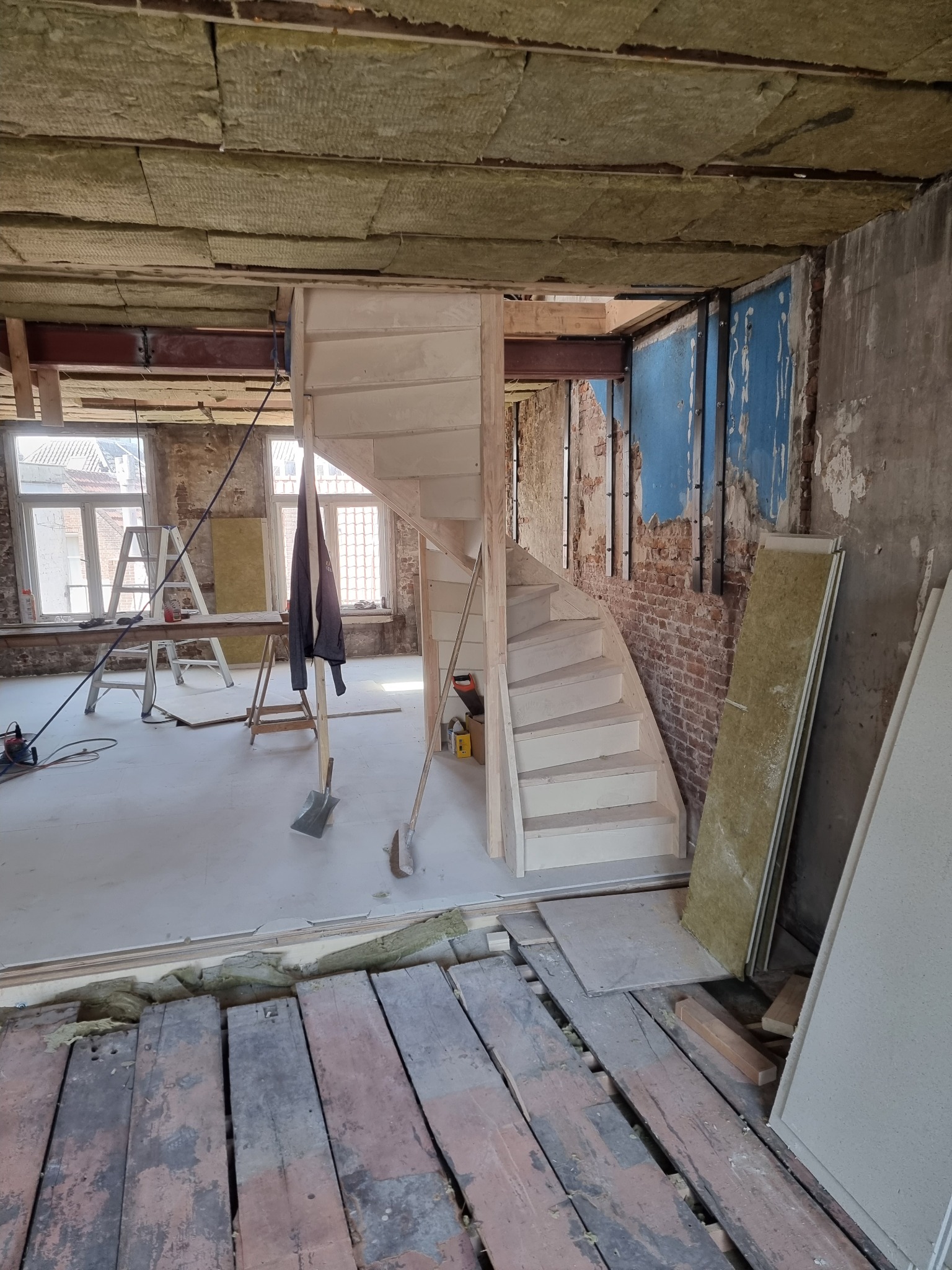 Wnętrze w trakcie prac, widoczne odsłonięte deski podłogowe, nowa drewniana konstrukcja schodów, izolacja na suficie, ceglana ściana z fragmentami niebieskiej farby, drabina i narzędzia.