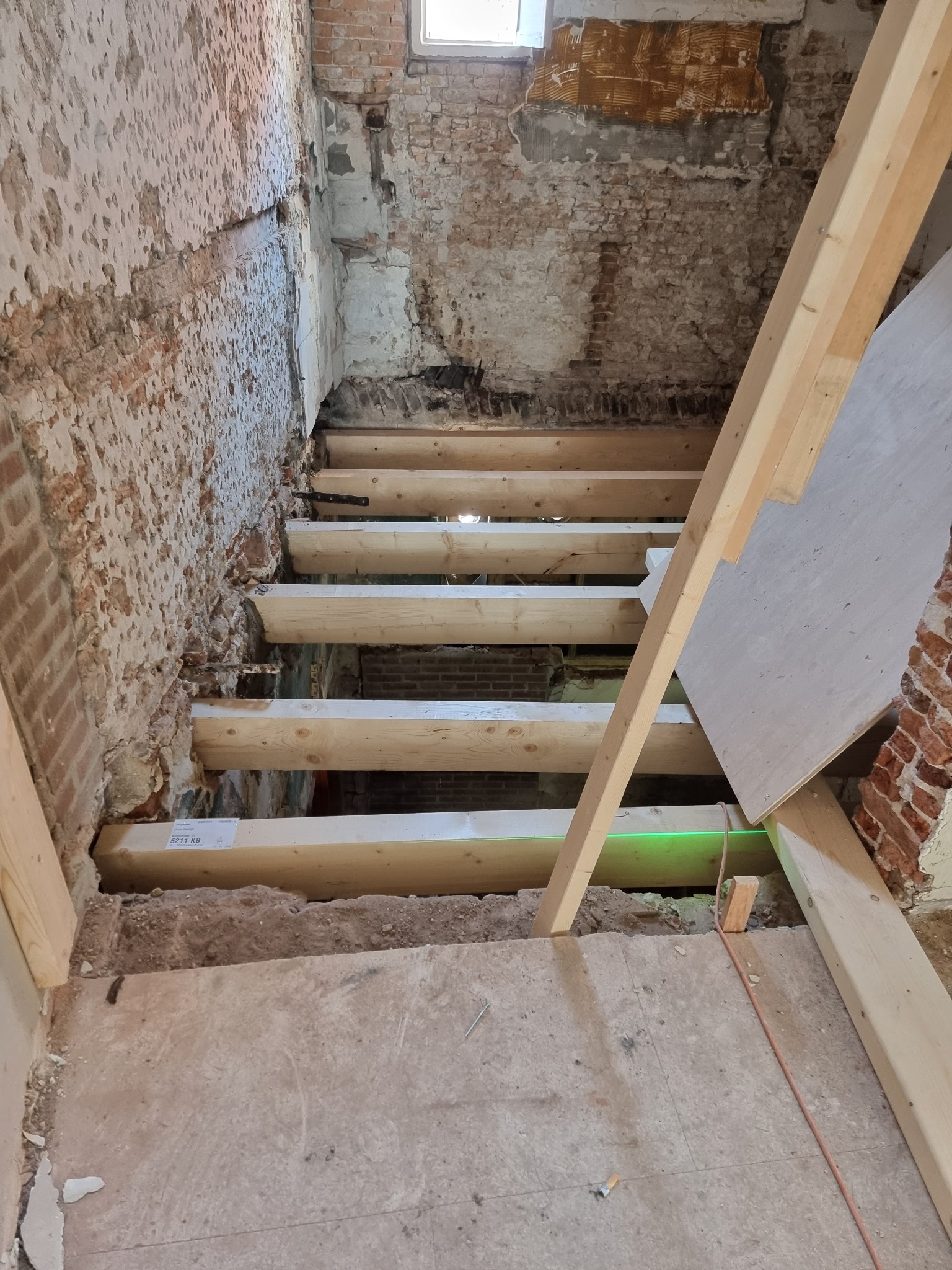 Konstrukcja schodów z surowego drewna w trakcie remontu, widoczne odsłonięte cegły i fragmenty starego tynku na ścianach, laserowy poziom wyznacza linię prostą, podłoże w stanie surowym.