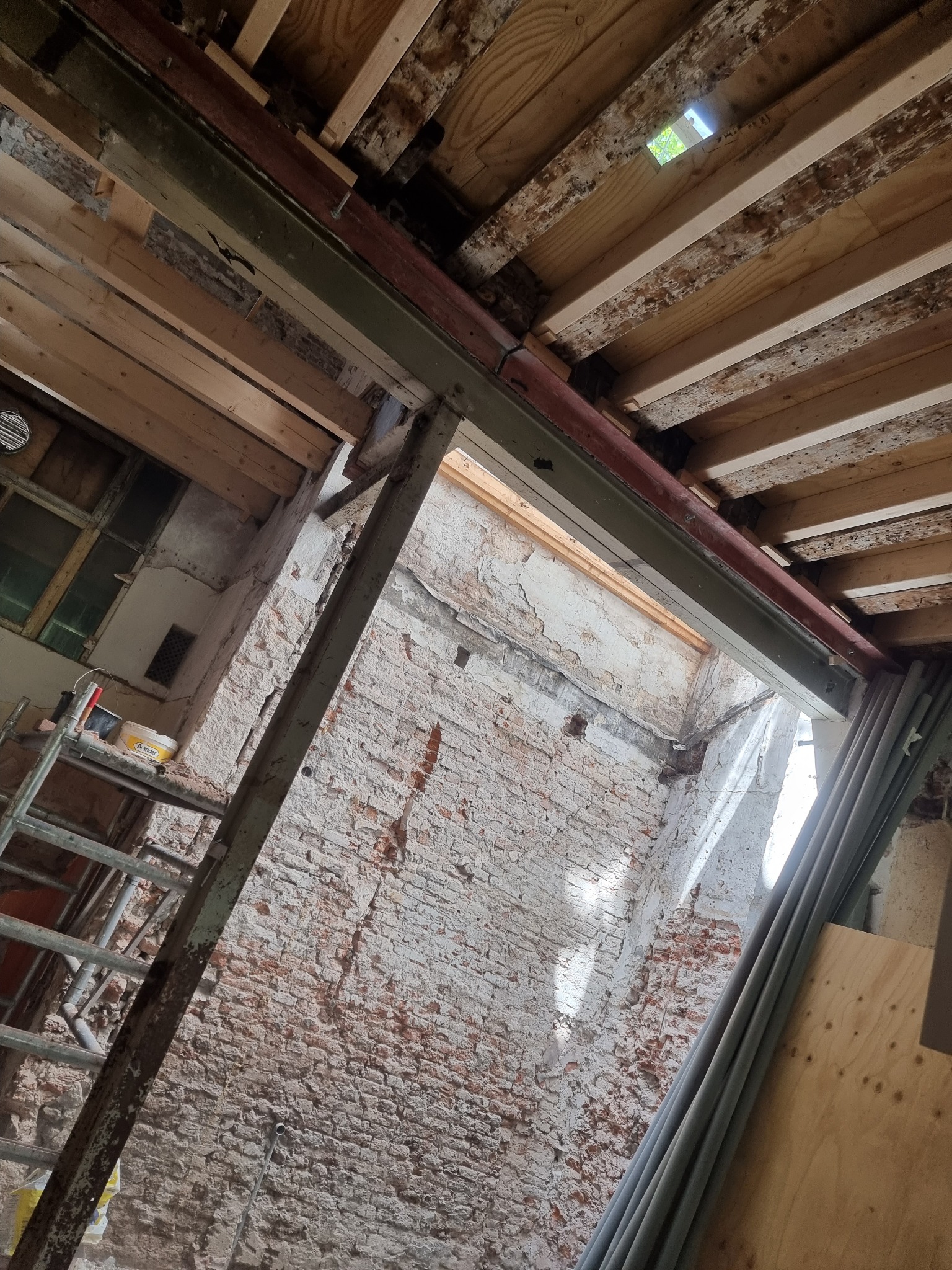 Wnętrze w trakcie gruntownego remontu: odsłonięta cegła, drewniane belki stropowe z widocznymi śladami wcześniejszych prac, metalowe wzmocnienia konstrukcyjne, drabina z narzędziami i puszką farby,...