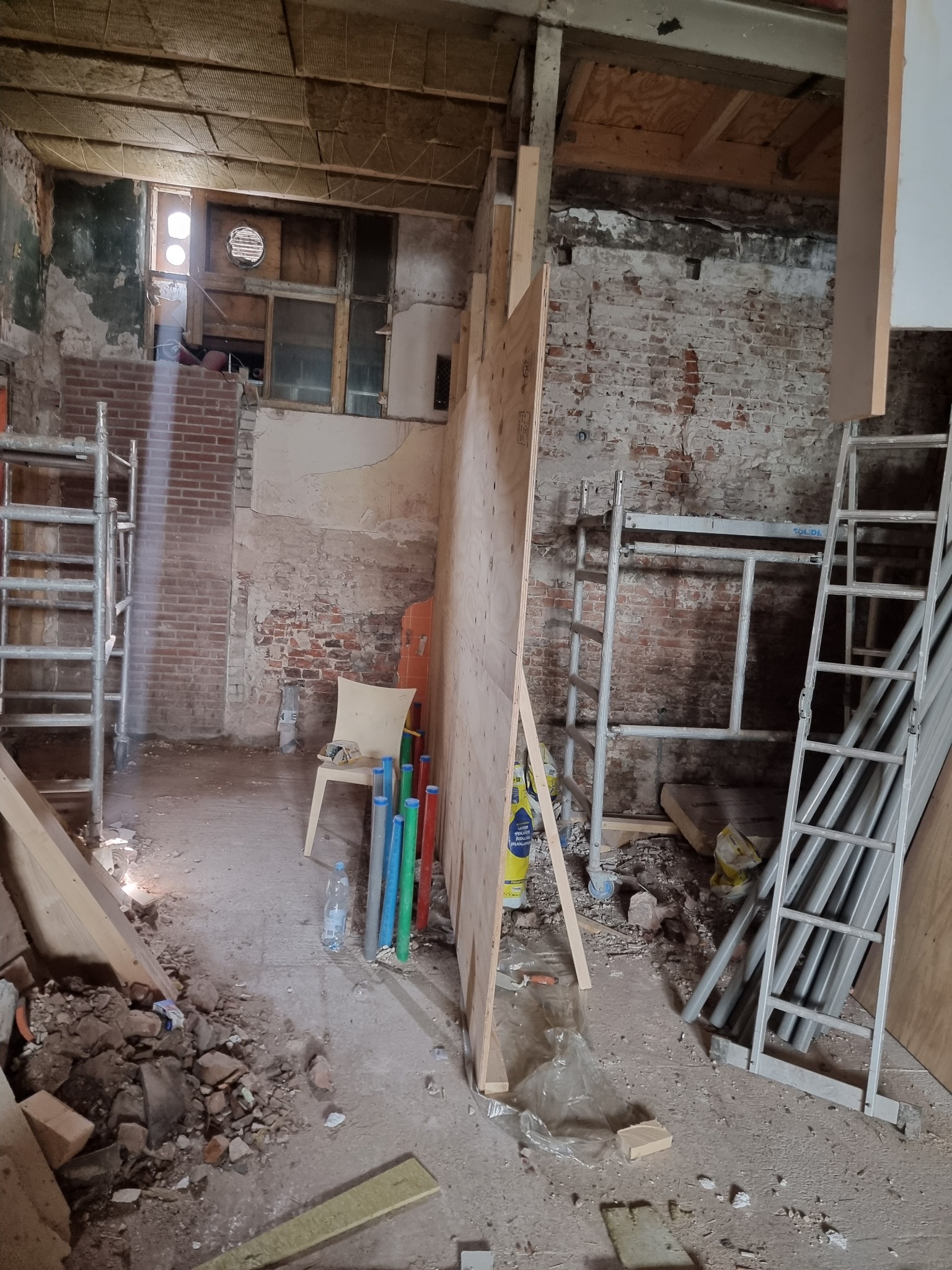 Wnętrze w stanie surowym z odsłoniętą cegłą, widoczne rusztowania, płyta OSB, rury PCV w kolorach zielonym, niebieskim i czerwonym, oraz krzesło. W tle okno z kratą i fragmenty instalacji.