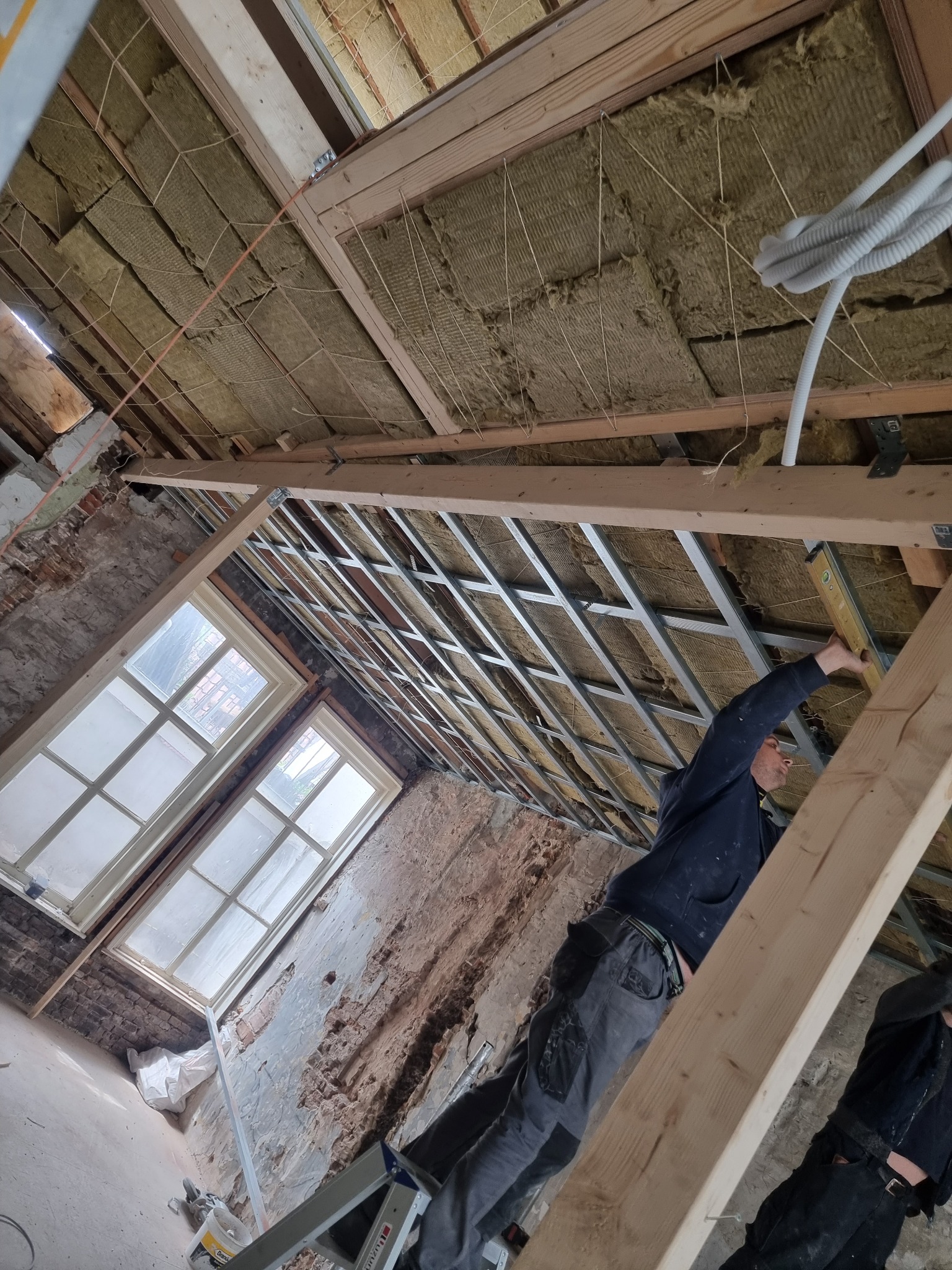 Montaż sufitu podwieszanego z wełną mineralną w pomieszczeniu z oknami, widoczny pracownik z poziomicą i fragmenty ściany z odsłoniętą cegłą.