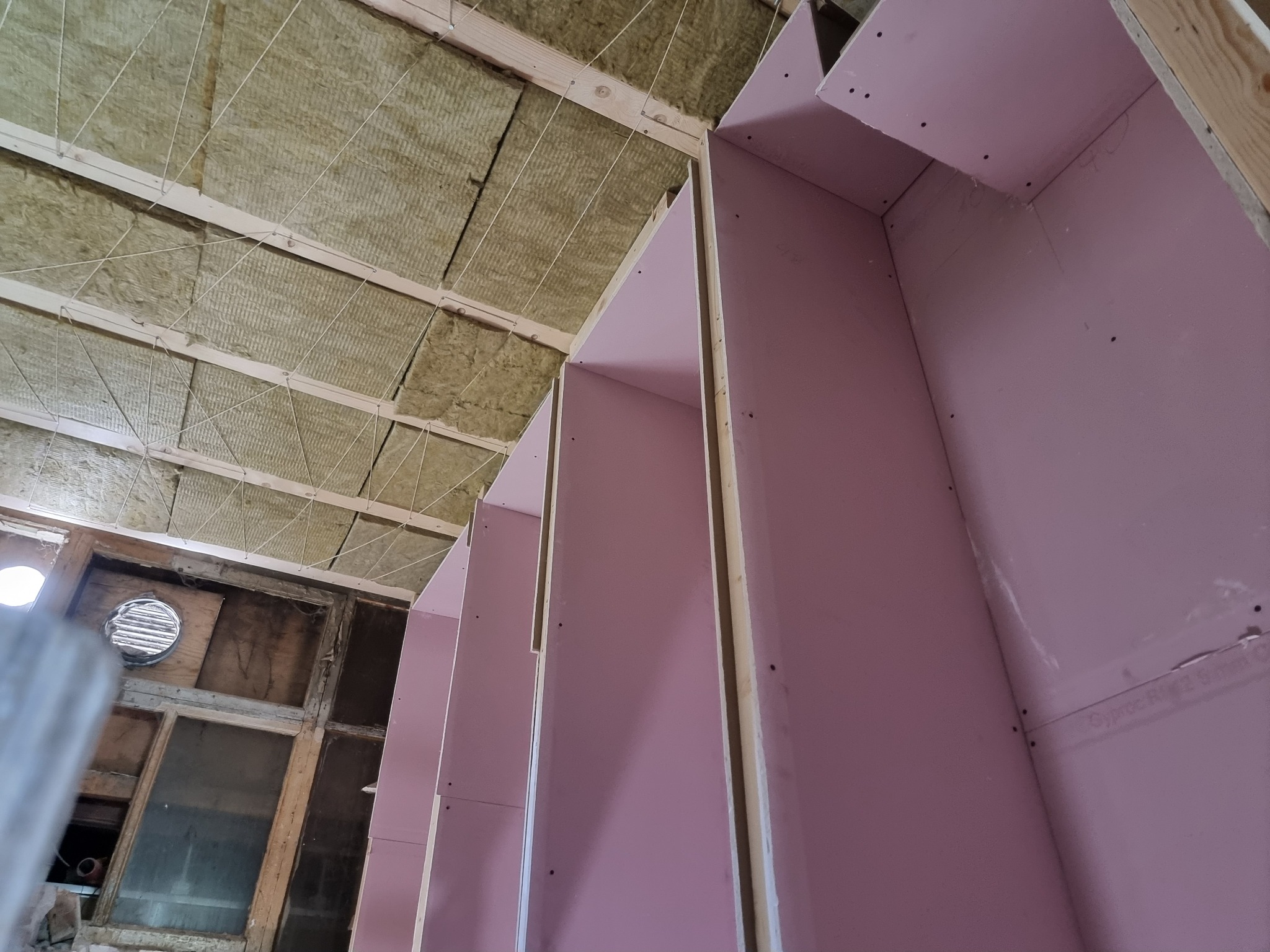 Wnętrze poddasza w trakcie prac wykończeniowych: izolacja wełną mineralną podwieszona sznurkami, montaż płyt gipsowo-kartonowych w kolorze różowym.