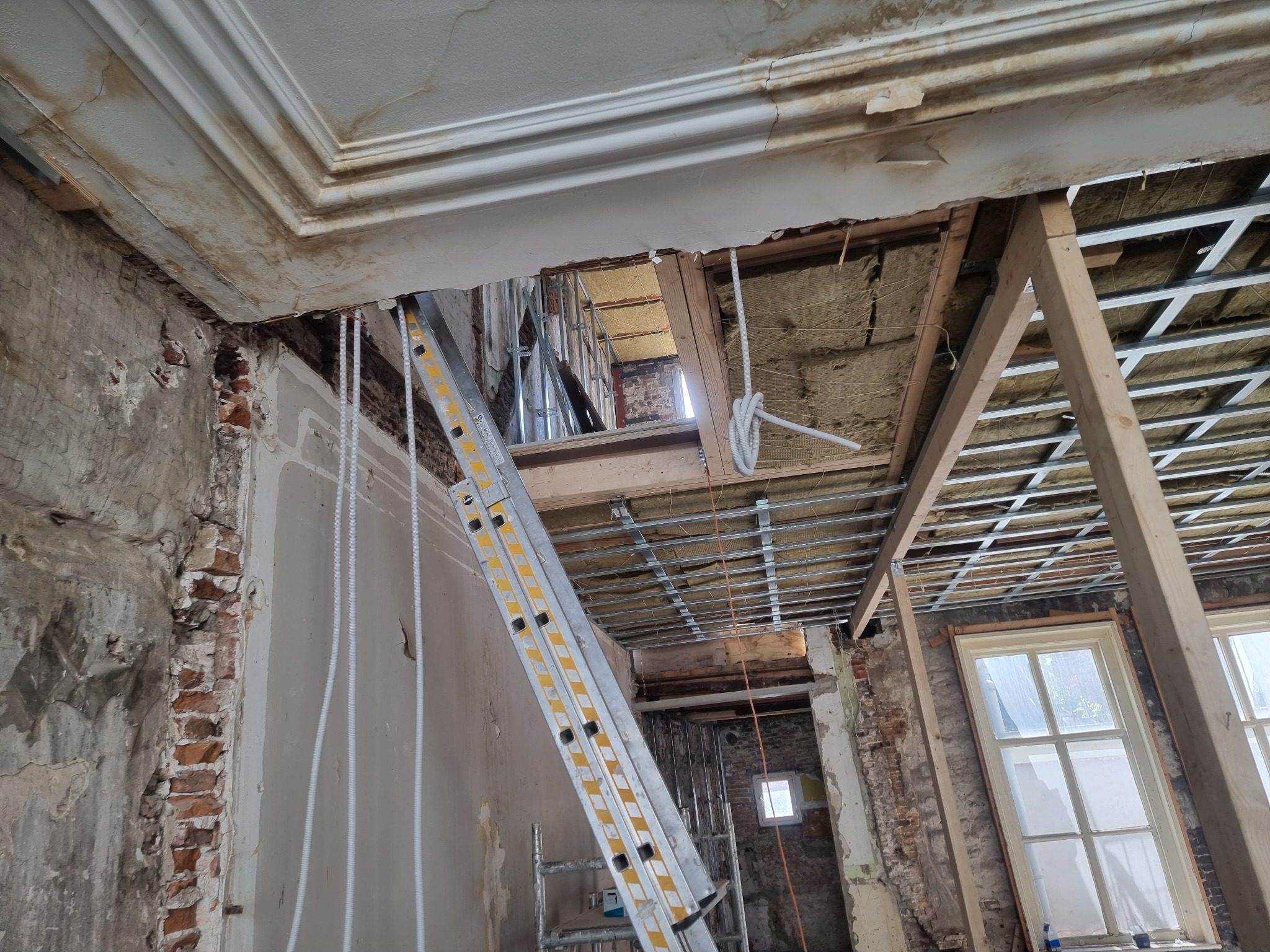 Remont mieszkania w Połczynie-Zdroju: widok na odsłonięty sufit z widoczną konstrukcją, izolacją i instalacjami elektrycznymi, drabina oparta o ścianę z fragmentami odsłoniętej cegły.