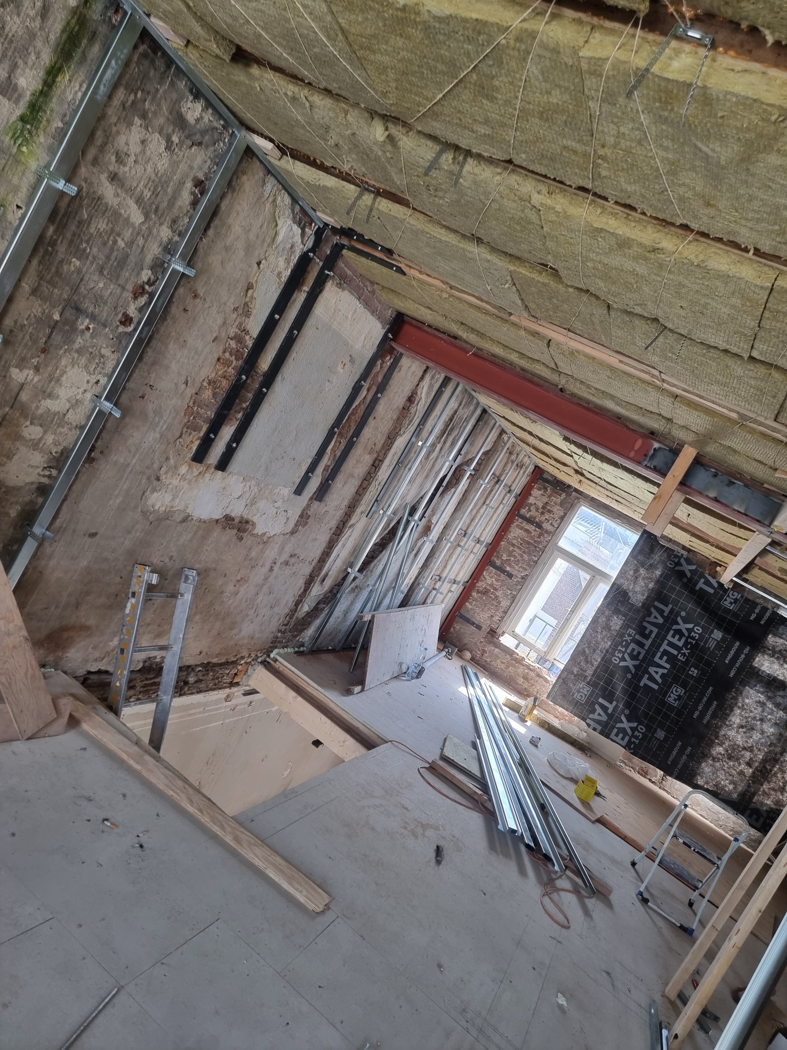 Poddasze w trakcie izolacji: widoczna wełna mineralna na suficie, metalowe profile ścian, częściowo położona podłoga, okno z widokiem na budynki, membrana TAFTEX na ścianie, drabina i narzędzia