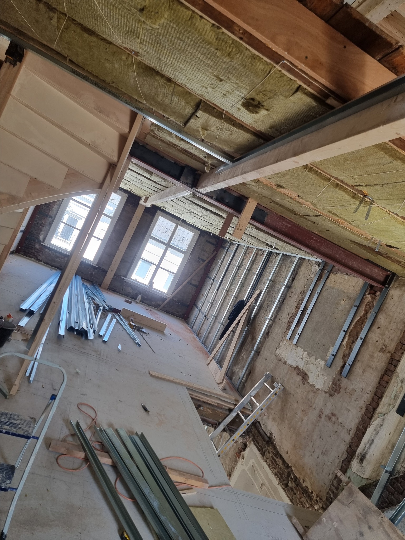 Wnętrze w trakcie prac: odsłonięta konstrukcja stropu z izolacją, metalowe profile ścienne, stare cegły, okna z widokiem na budynek, rozłożone materiały i narzędzia na podłodze.