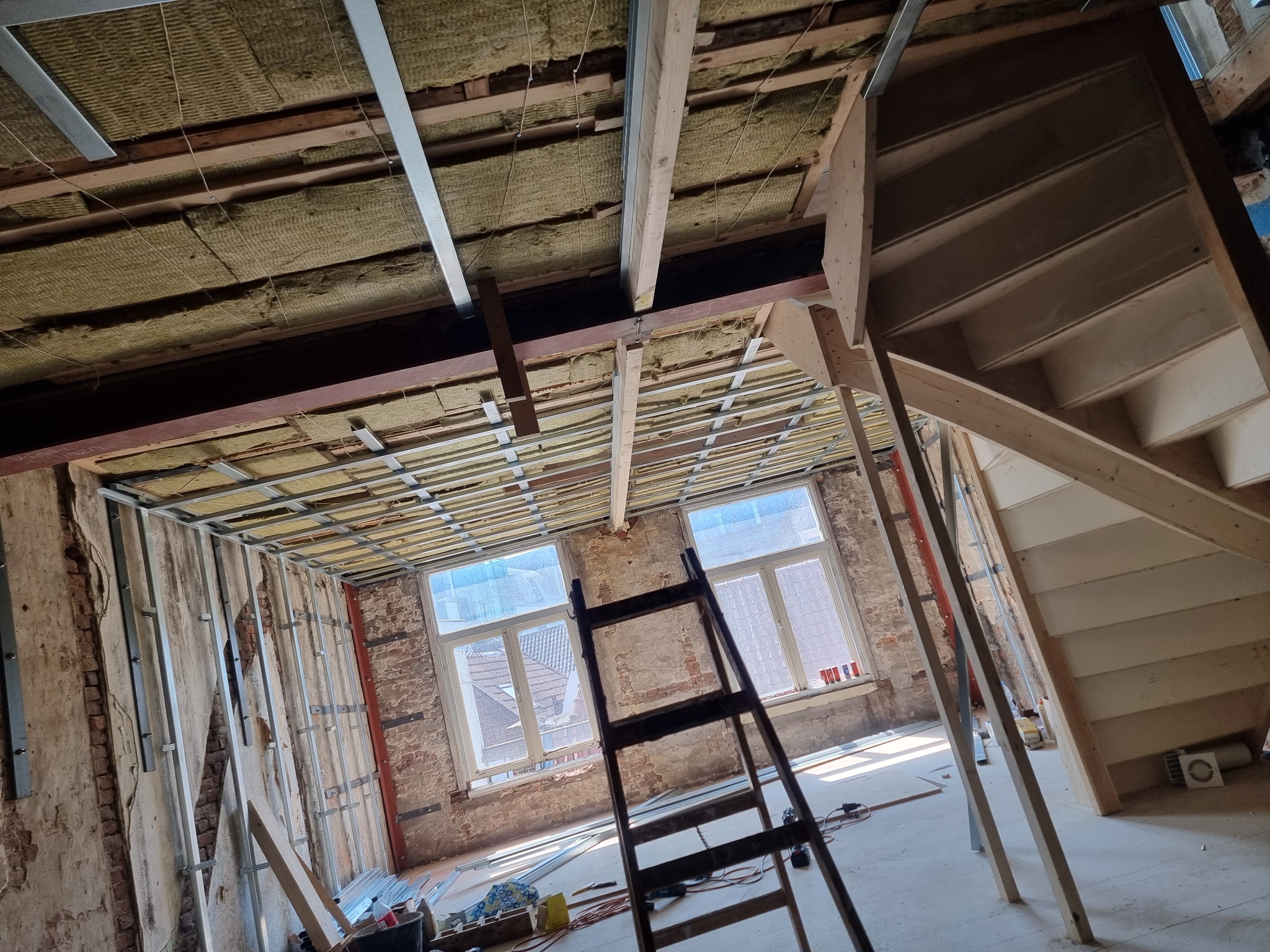 Wnętrze w trakcie prac: izolacja stropu wełną mineralną, metalowy stelaż pod sufitem, surowe ściany z cegły, drewniana konstrukcja schodów, drabina oparta o ścianę.