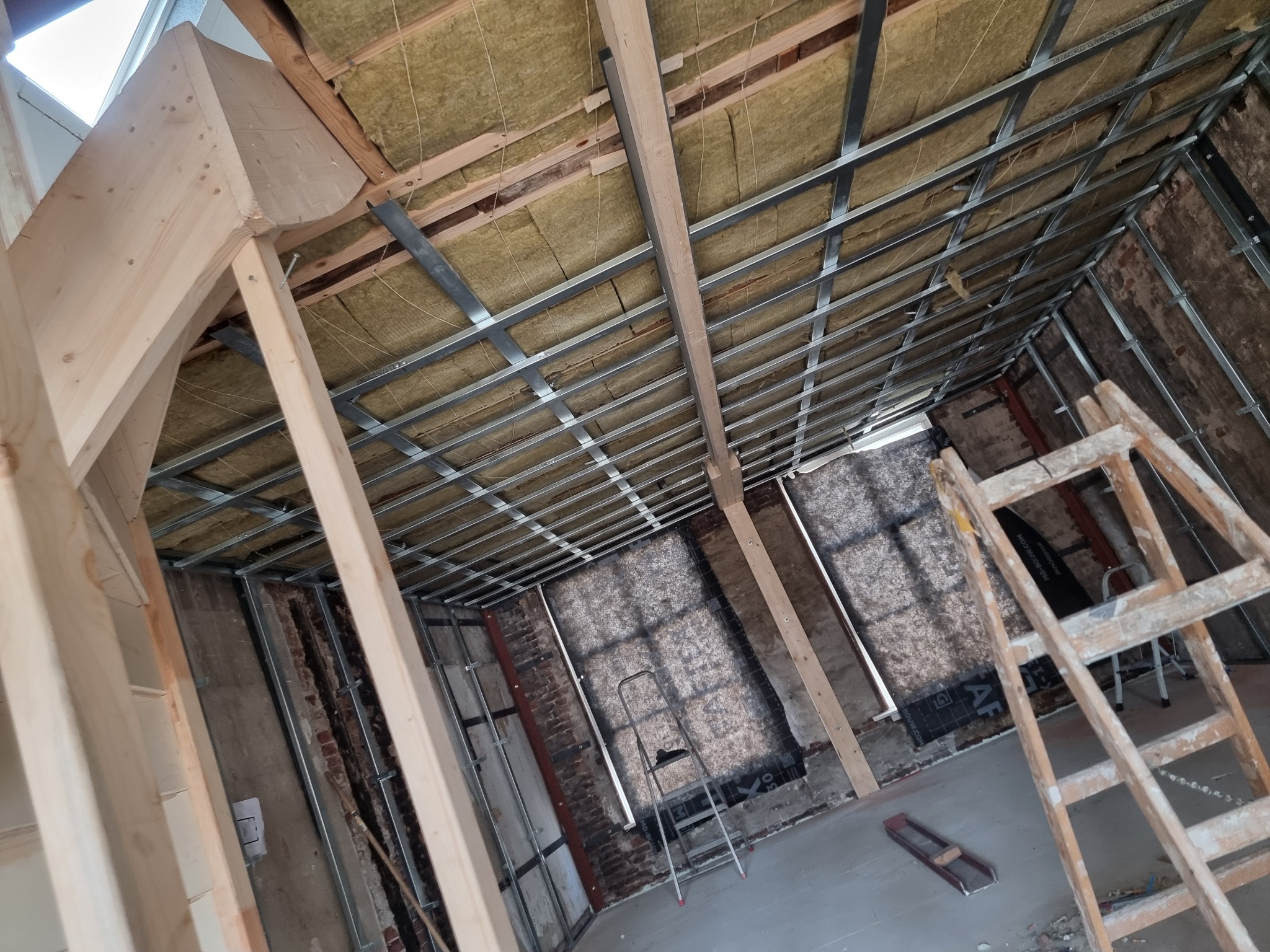 Wnętrze w trakcie prac: montaż metalowego stelaża sufitu podwieszanego z widoczną izolacją z wełny mineralnej i elementami drewnianej konstrukcji oraz drabina oparta o ścianę z fragmentami starej...