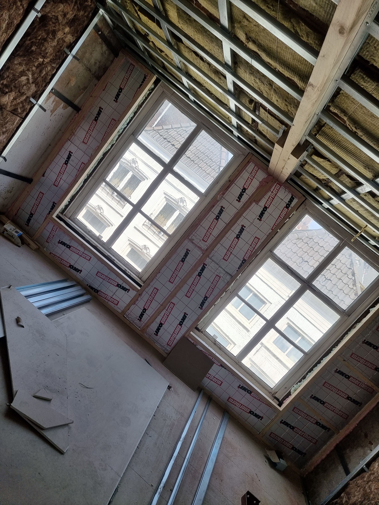 Montaż izolacji termicznej wokół okien dachowych w trakcie poddasza, widoczne profile metalowe konstrukcji sufitu i wełna mineralna.