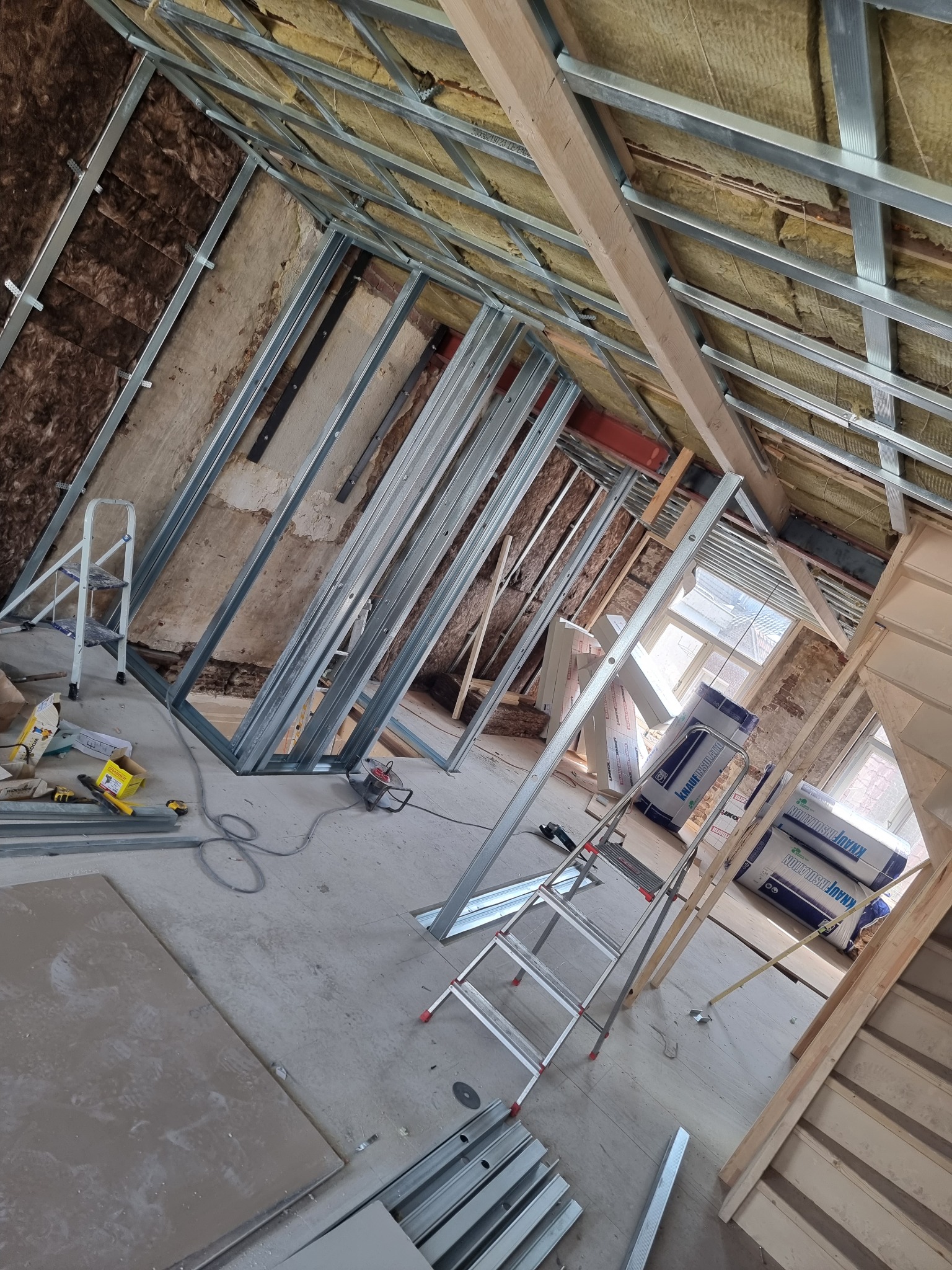 Montaż metalowej konstrukcji pod sufit napinany na poddaszu, widoczna izolacja wełną mineralną, drabina i narzędzia na podłodze.