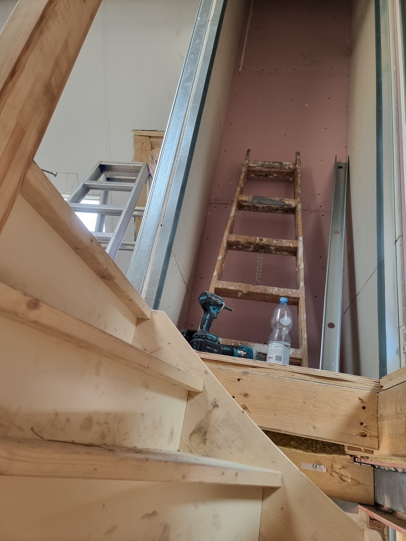 Montaż drewnianych schodów w surowym stanie, widoczne narzędzia, drabina i płyta gipsowa w tle.