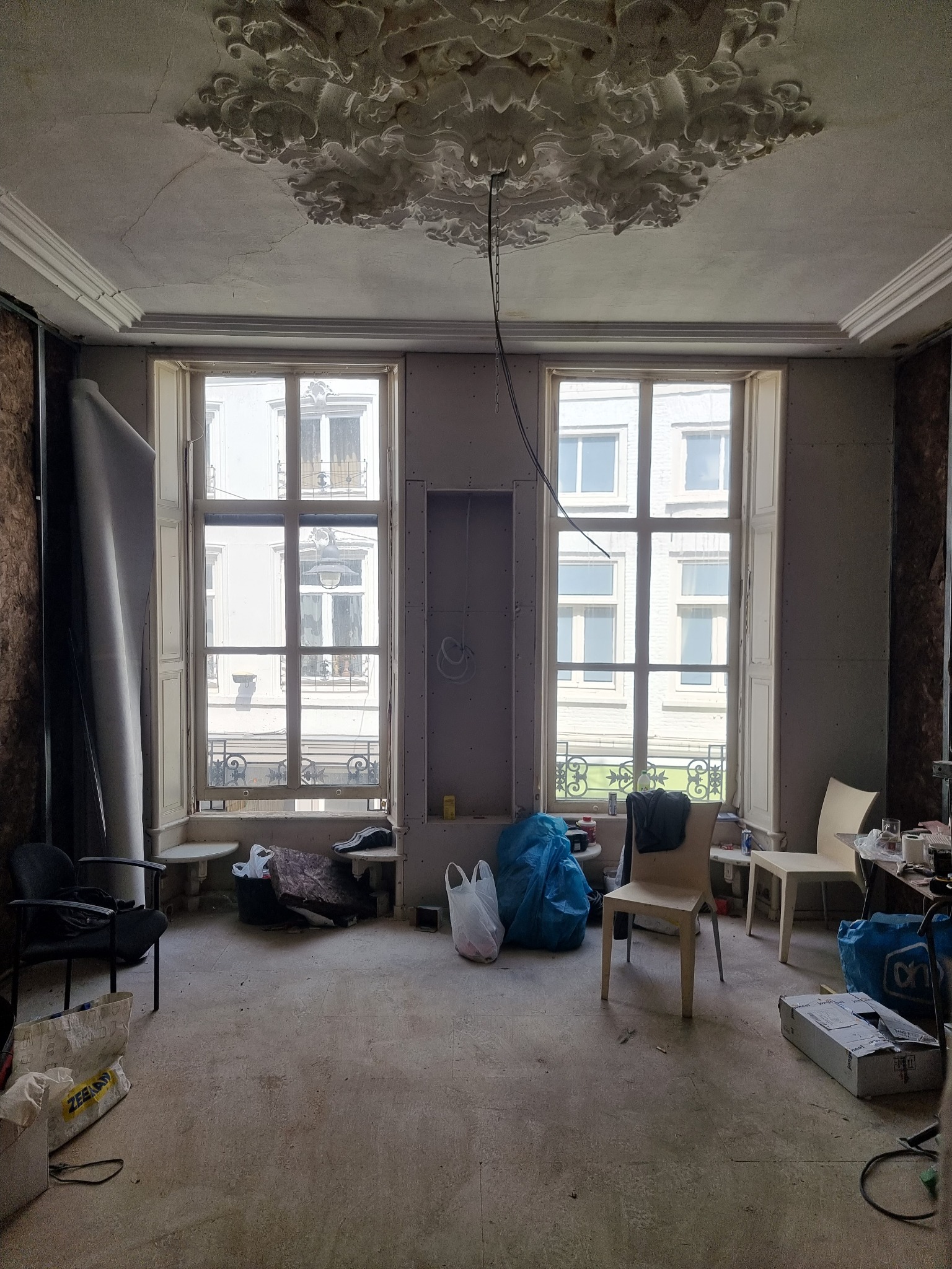 Pokój w trakcie przygotowania do prac wykończeniowych, widoczne dwie duże okna z widokiem na budynek naprzeciwko, ściany przygotowane do malowania, ozdobny gipsowy sufit z widocznymi pęknięciami...