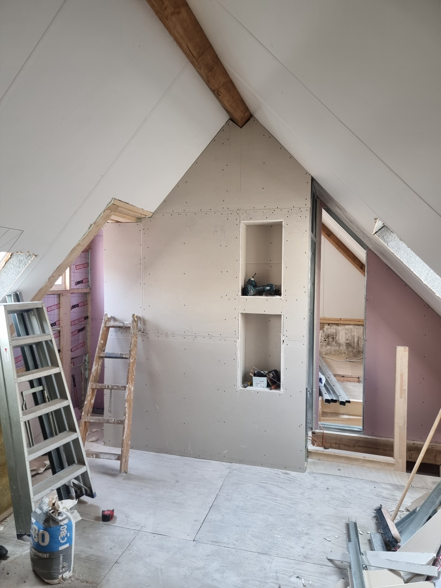 Poddasze w trakcie adaptacji: ściany z płyt kartonowo-gipsowych z wnękami, widoczne ocieplenie, dwie drabiny, worek cementu i elementy konstrukcyjne na podłodze.