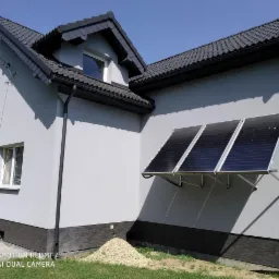 Ocieplony szary dom jednorodzinny z ciemną dachówką i trzema panelami słonecznymi zamontowanymi na elewacji bocznej, widoczny fragment trawnika i kupka kamieni.