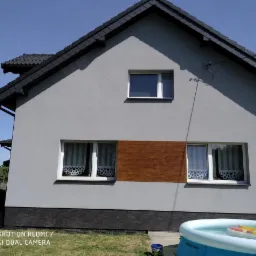 Szary dom jednorodzinny z ciemną dachówką i brązowym panelem elewacyjnym między oknami, widoczny basen ogrodowy.