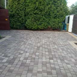 Ułożona kostka brukowa w odcieniach szarości i beżu, otoczona zielenią iglaków i elementami ogrodu, widoczne niedokończone obrzeże z kostki.