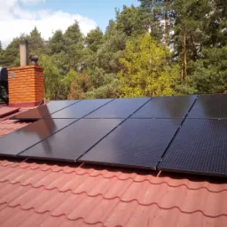 Czarny panel słoneczny na czerwonym dachu z blachodachówki, widoczny komin z cegły i zbiornik solarny, w tle drzewa iglaste i liściaste.