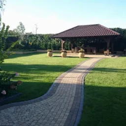 Ułożona ścieżka z jasnobrązowej kostki prowadzi przez zadbany trawnik do drewnianej altany z ciemnym dachem, w tle żywopłot i murowany grill.