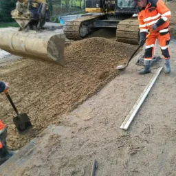 Przygotowanie podłoża pod nawierzchnię: robotnicy rozprowadzają żwir koparką i łopatami, widoczne obrzeża chodnika.