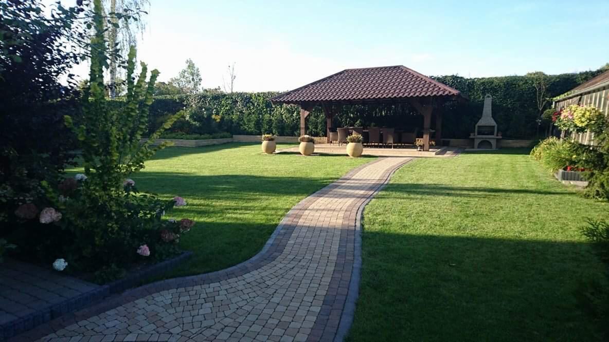 Ułożona ścieżka z jasnobrązowej kostki prowadzi przez zadbany trawnik do drewnianej altany z ciemnym dachem, w tle żywopłot i murowany grill.