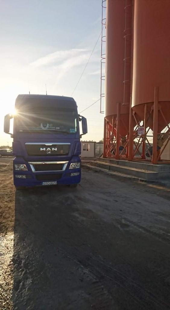 Niebieska ciężarówka MAN na tle silosów do przechowywania materiałów sypkich, w tle widoczny budynek biurowy, wczesne poranne słońce.