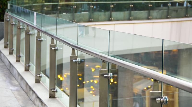 Szklana balustrada z metalowymi słupkami i poręczą, widok z boku na tle rozmytych świateł.
