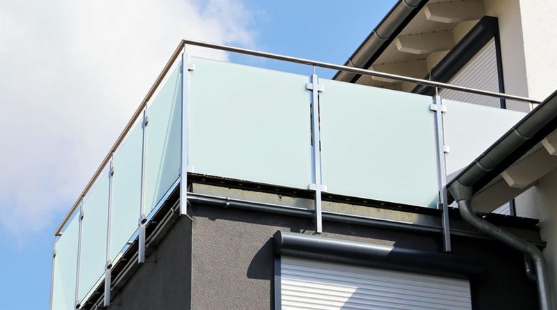 Nowoczesna balustrada balkonowa z matowego szkła i stali nierdzewnej, widok z dołu na tle błękitnego nieba.