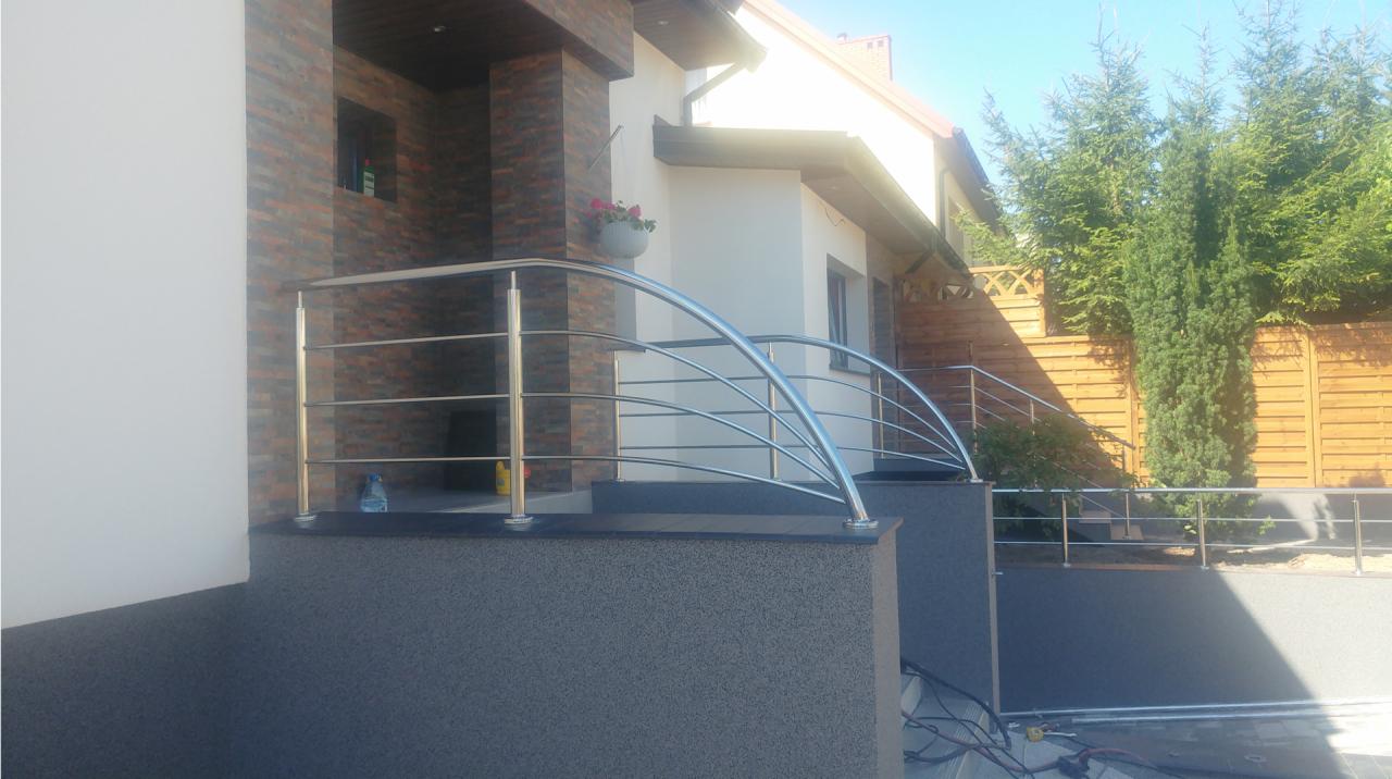 Zewnętrzne schody z ciemnego kamienia z nowoczesną balustradą ze stali nierdzewnej, prowadzące do wejścia domu z ceglaną elewacją i kwiatami w doniczce.