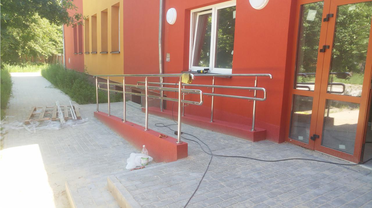 Zewnętrzna rampa dla niepełnosprawnych z balustradą ze stali nierdzewnej przy wejściu do budynku o pomarańczowo-żółtej elewacji, widoczne narzędzia i pozostałości po montażu.