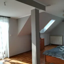 Pokój na poddaszu po remoncie, ściany pomalowane na szaro, drewniana podłoga, okno dachowe, widoczna kolumna konstrukcyjna i fragment łóżka.