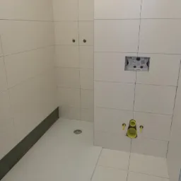 Biała, kwadratowa kabina prysznicowa bez drzwi, z odpływem liniowym w podłodze i białymi płytkami na ścianach, widoczne instalacje wodno-kanalizacyjne w trakcie montażu.