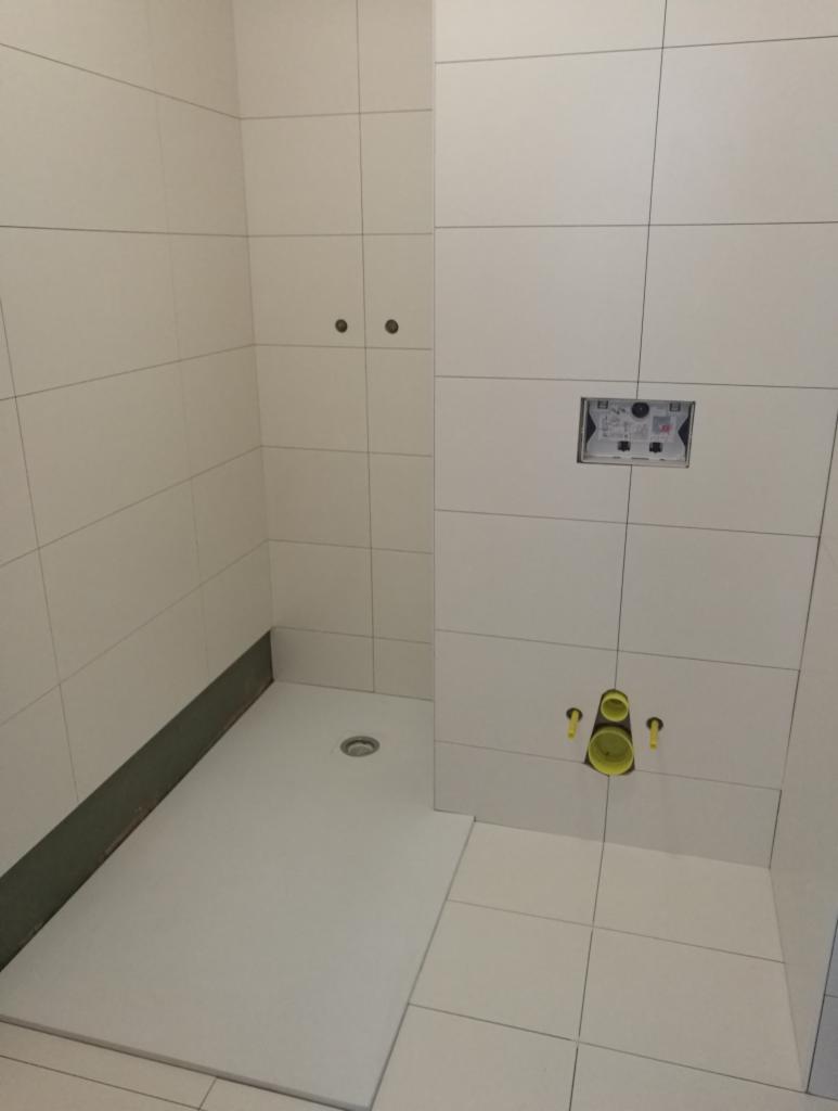 Biała, kwadratowa kabina prysznicowa bez drzwi, z odpływem liniowym w podłodze i białymi płytkami na ścianach, widoczne instalacje wodno-kanalizacyjne w trakcie montażu.