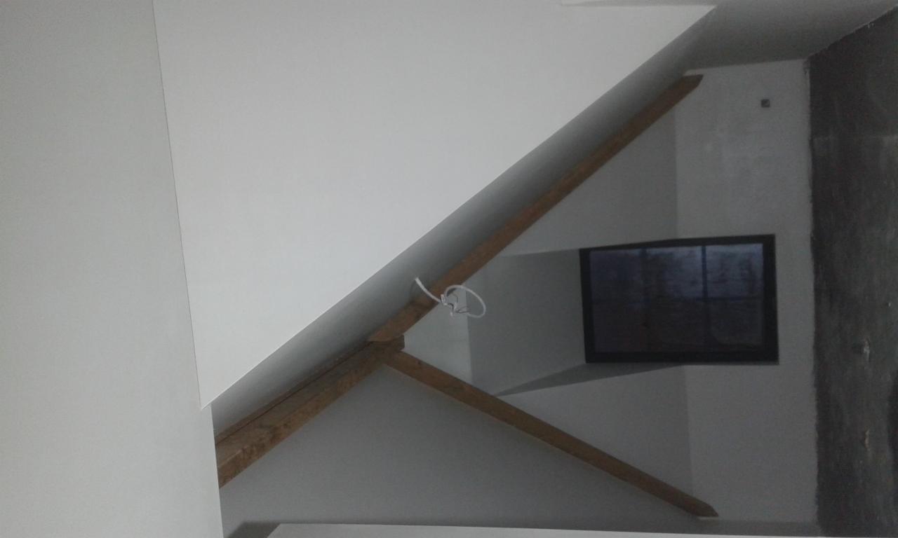 Poddasze po remoncie z widocznymi drewnianymi belkami konstrukcyjnymi, świeżo pomalowane ściany na biało i okno w czarnej ramie.