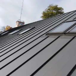 Nowoczesne, grafitowe pokrycie dachowe z blachy na dachu skośnym z oknami dachowymi, widok z dołu, w tle budynek z wieżą telekomunikacyjną.