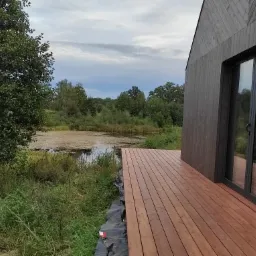 Nowoczesny taras drewniany przylegający do ciemnej elewacji domu, z widokiem na zarośnięty staw i las w tle.