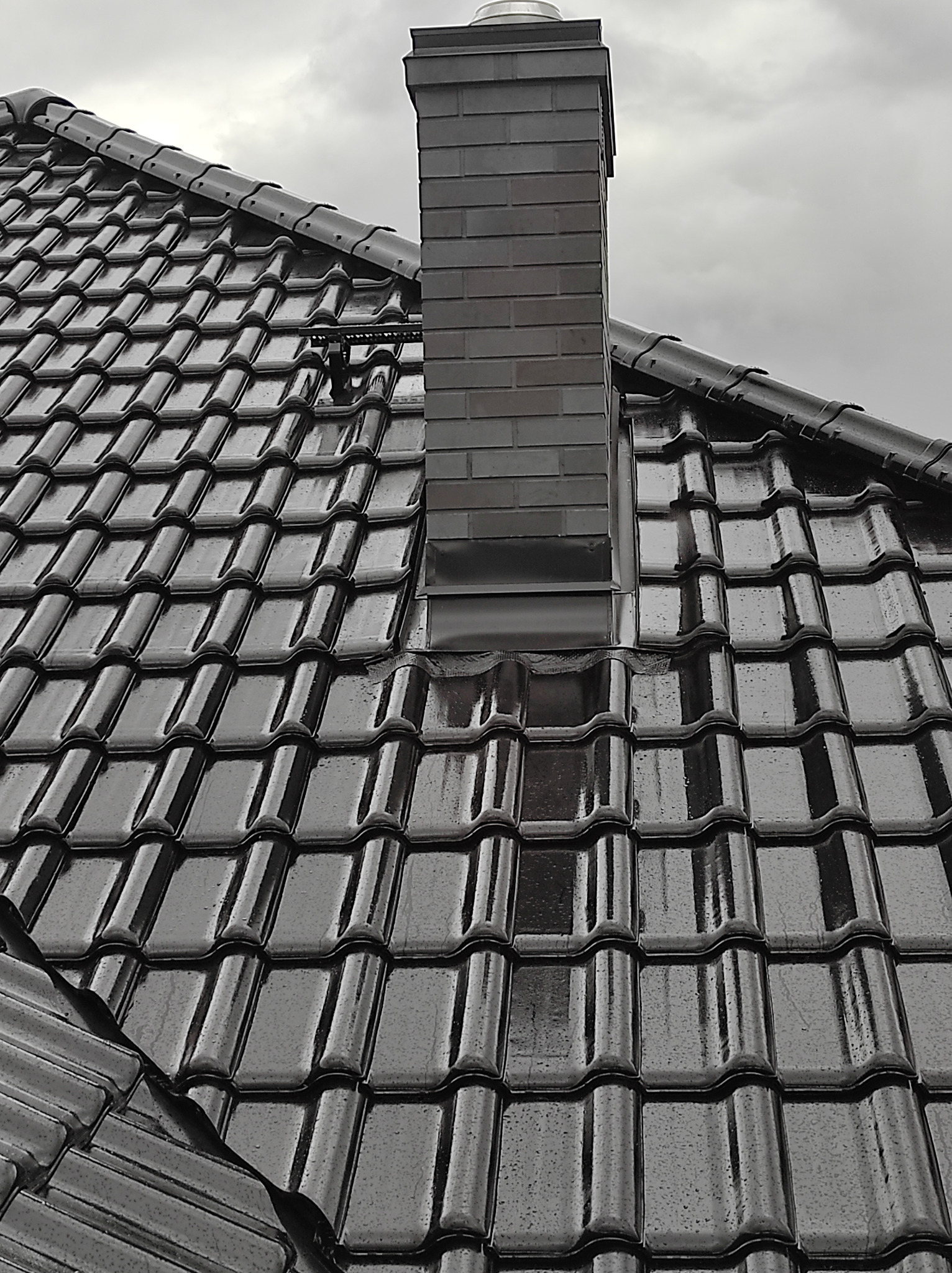 Błyszcząca, mokra od deszczu dachówka ceramiczna w kolorze grafitowym, z widocznym kominem z cegły klinkierowej i zabezpieczeniem przeciwśniegowym.