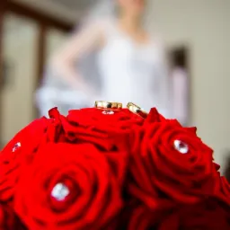 Dwa złote obrączki ślubne spoczywają na bukiecie czerwonych róż zdobionych kryształkami, w tle rozmyta sylwetka panny młodej w białej sukni.