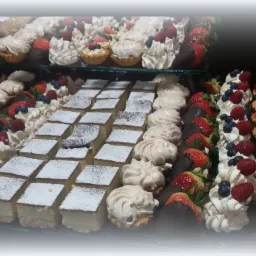 Różnorodny wybór ciast i deserów na tacy: kwadratowe ciasta posypane cukrem pudrem, mini bezy, tartaletki z kremem i owocami (truskawki, maliny, borówki), truskawki w czekoladzie i kanapki.