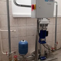 Zmontowana instalacja hydroforowa z niebieskim zbiornikiem ciśnieniowym, pompą pionową i skrzynką sterowniczą z wyłącznikiem, na tle beżowych płytek ceramicznych.