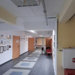 Korytarz biurowy z widocznymi rurami instalacyjnymi pod sufitem, skrzynką hydrantową na ścianie i dyplomami na ścianie po lewej stronie.