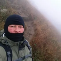 Mężczyzna w kominiarce i kurtce, z plecakiem, na szlaku górskim we mgle. Widoczny fragment ścieżki i trawa. Brak widoczności w oddali.