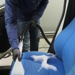 Mężczyzna w czarnych rękawicach i lampą czołową czyści niebieskie siedzenie samochodowe za pomocą odkurzacza piorącego, pozostawiając ślady piany.