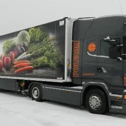 Oklejona ciężarówka firmy Polfarm z grafiką warzyw na naczepie, stojąca na ośnieżonym terenie w pochmurny dzień.