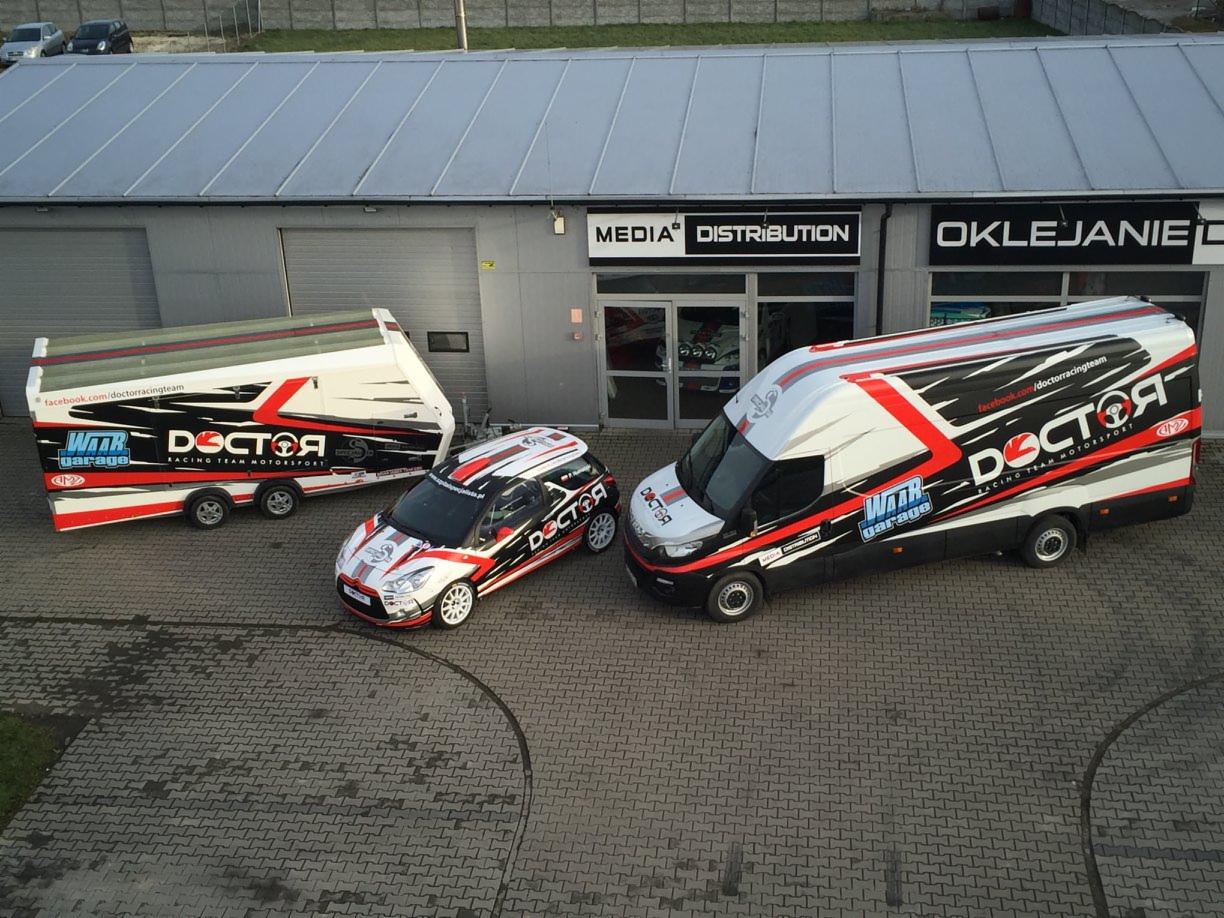 Zestaw pojazdów rajdowych - samochód osobowy, bus i przyczepa - z identycznym brandingiem Doctor Racing Team Motorsport, zaparkowane przed budynkiem z napisem OKLEJANIE.