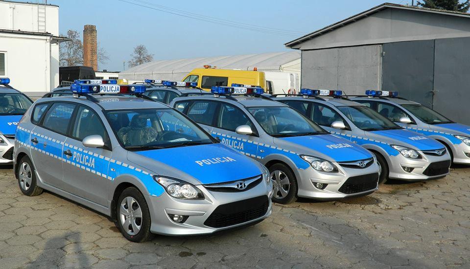 Rząd srebrnych samochodów policyjnych Hyundai i30 z niebieskimi pasami i napisami 'Policja', ustawionych na zewnątrz budynku w słoneczny dzień.