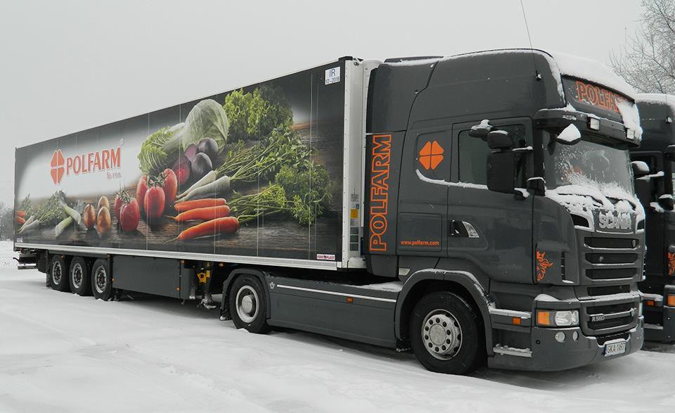 Oklejona ciężarówka firmy Polfarm z grafiką warzyw na naczepie, stojąca na ośnieżonym terenie w pochmurny dzień.